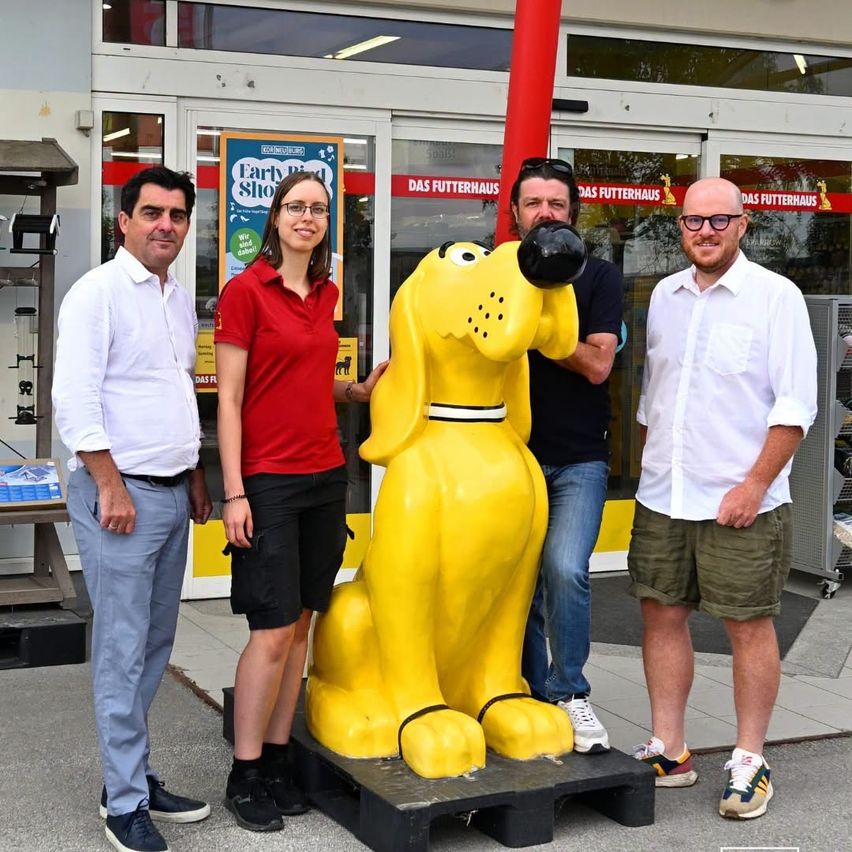 Vier Personen stehen vor einer großen gelben Hundestatue und lächeln für ein Foto. Dahinter befindet sich ein Geschäft mit dem Schild 'Das Futterhaus.'