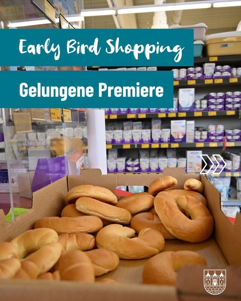 Eine Schachtel mit verschiedenen Broten, darunter Brezeln und Brötchen, wird in einem Geschäft mit Regalen voller Produkte im Hintergrund ausgestellt. Das Bild hat einen Wasserzeichen mit dem Text 'Early Bird Shopping' und 'Gelungene Premiere'.