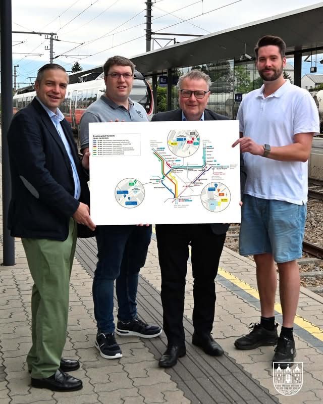 Vier Männer posieren mit einer Karte am Bahnhof und halten eine Karte mit Zugverbindungen. Hinter ihnen ist ein Zug zu sehen.