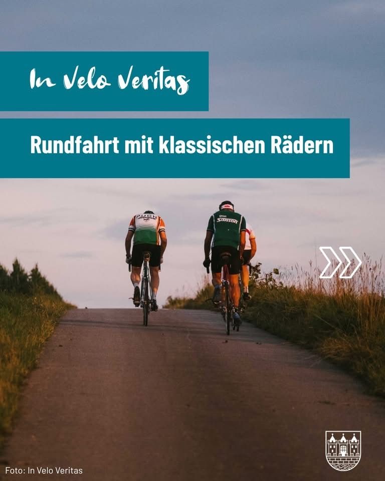 Zwei Radfahrer fahren auf einer Straße mit klassischen Fahrrädern. Der Hintergrund zeigt einen bewölkten Himmel und ein Feld mit hohem Gras.