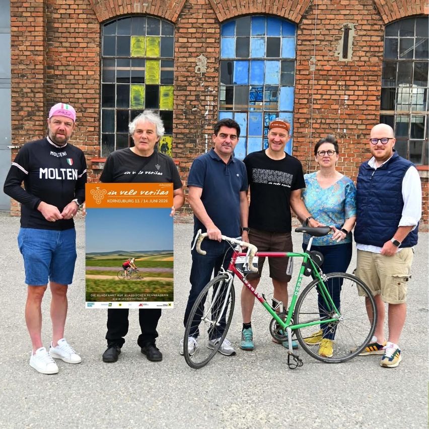 Eine Gruppe von Menschen posiert mit Fahrrädern vor einem Backsteingebäude. Sie tragen Freizeitkleidung. Ein Mann hält ein Plakat mit einem Fahrrad darauf.