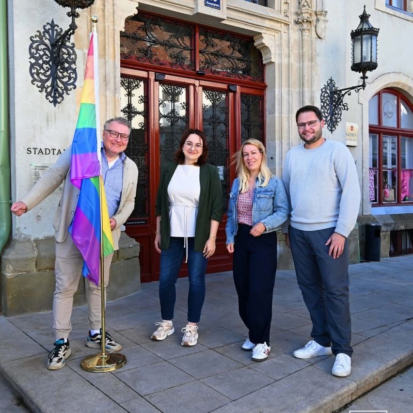 Vier Personen stehen vor einem Gebäude mit einer Regenbogenflagge und lächeln für ein Foto. Der Mann auf der linken Seite hält die Flagge. Sie sind lässig gekleidet, zwei tragen Brillen. Das Gebäude hat eine rote Tür und aufwendige Verzierungen.