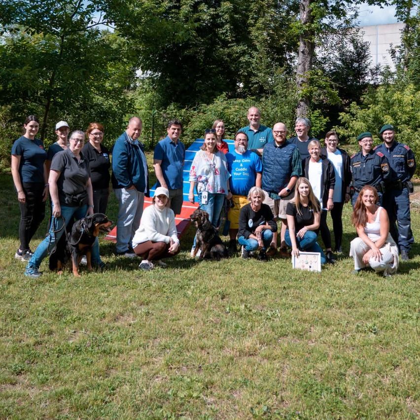 Eine Gruppe von Menschen und Hunden posiert für ein Foto auf einer Wiese, einige stehend, andere kniend. Bäume und ein Gebäude sind im Hintergrund.