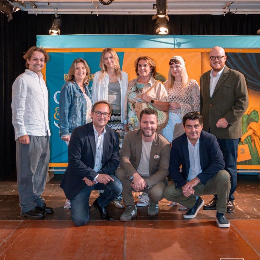 Eine Gruppe von Menschen posiert für ein Foto auf einer Bühne mit blauem und orangefarbenem Hintergrund. Sie lächeln und scheinen Teil eines Teams zu sein.