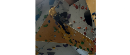 Eine Person in einem gelben Shirt und schwarzen Hosen klettert an einer mehrfarbigen Wand mit verschiedenen Griffen und Haltepunkten.