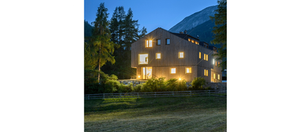 Ein modernes, beleuchtetes Haus steht in einer ruhigen Bergumgebung mit Bäumen und einem Berg im Hintergrund.