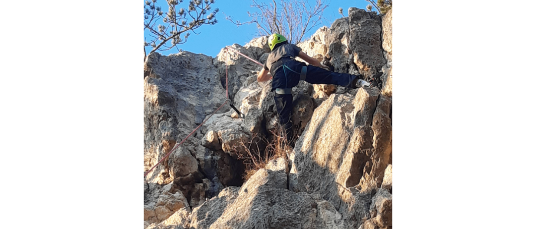 Eine Person klettert mit einem Seil an einem felsigen Felsen und trägt einen grünen Helm und einen Gurt, unter einem klaren blauen Himmel.