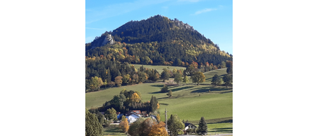 Ein Berg mit einem felsigen Gipfel ist von einem üppigen grünen Tal mit Bäumen in verschiedenen Farben umgeben. Ein paar Häuser sind in das Tal eingebettet.