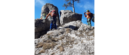 Drei Kletterer steigen einen felsigen Berg hinauf, einer macht ein Foto, während die anderen mit Helmen und Rucksäcken ausgestattet sind.