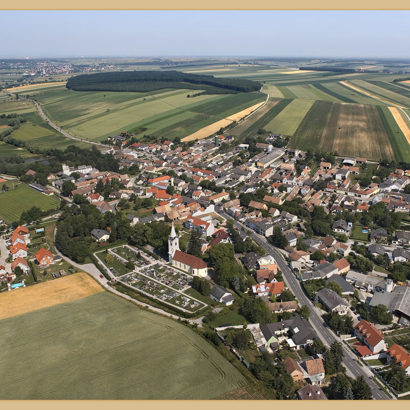 Luftaufnahme eines Dorfes mit vielen Häusern, einer Kirche und großen Feldern, die es umgeben. Das Dorf hat eine Mischung aus modernen und traditionellen Gebäuden, von denen einige rote Dächer haben. Die Felder sind weitläufig und scheinen Teil eines größeren landwirtschaftlichen Gebiets zu sein.