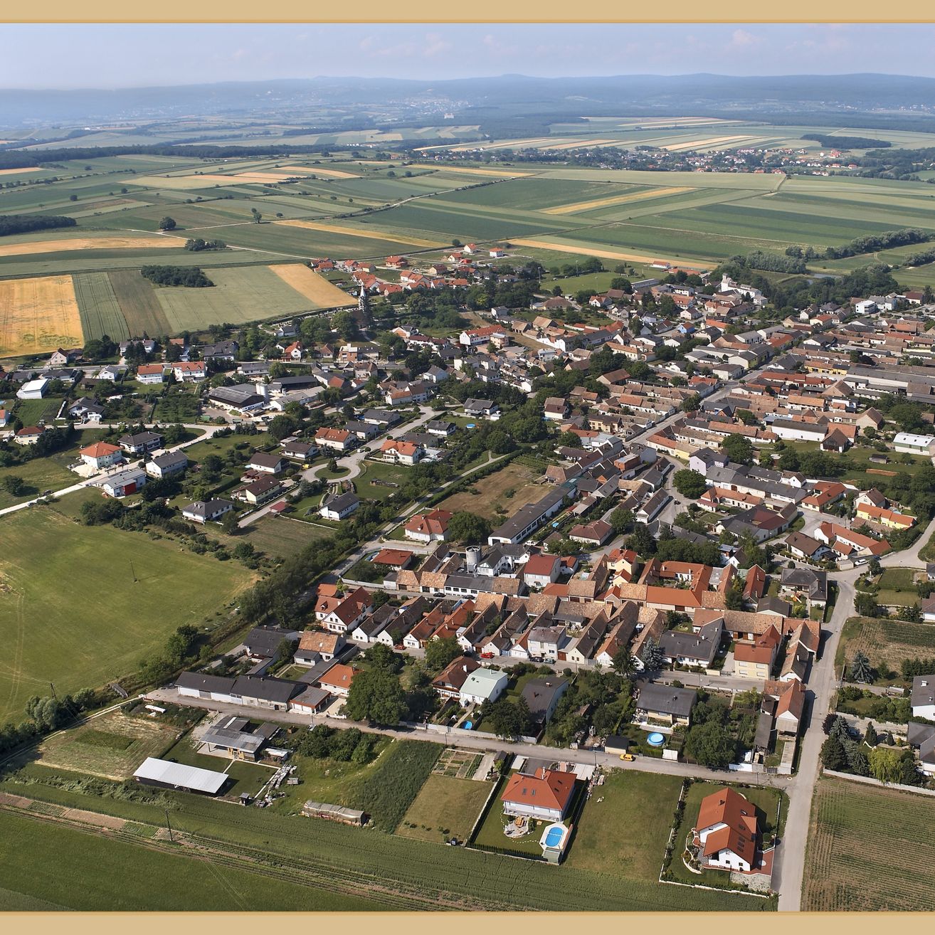 Eine Luftaufnahme eines ländlichen Dorfes mit zahlreichen Häusern, umgeben von grünen Feldern und Bäumen. Berge sind in der Ferne sichtbar.