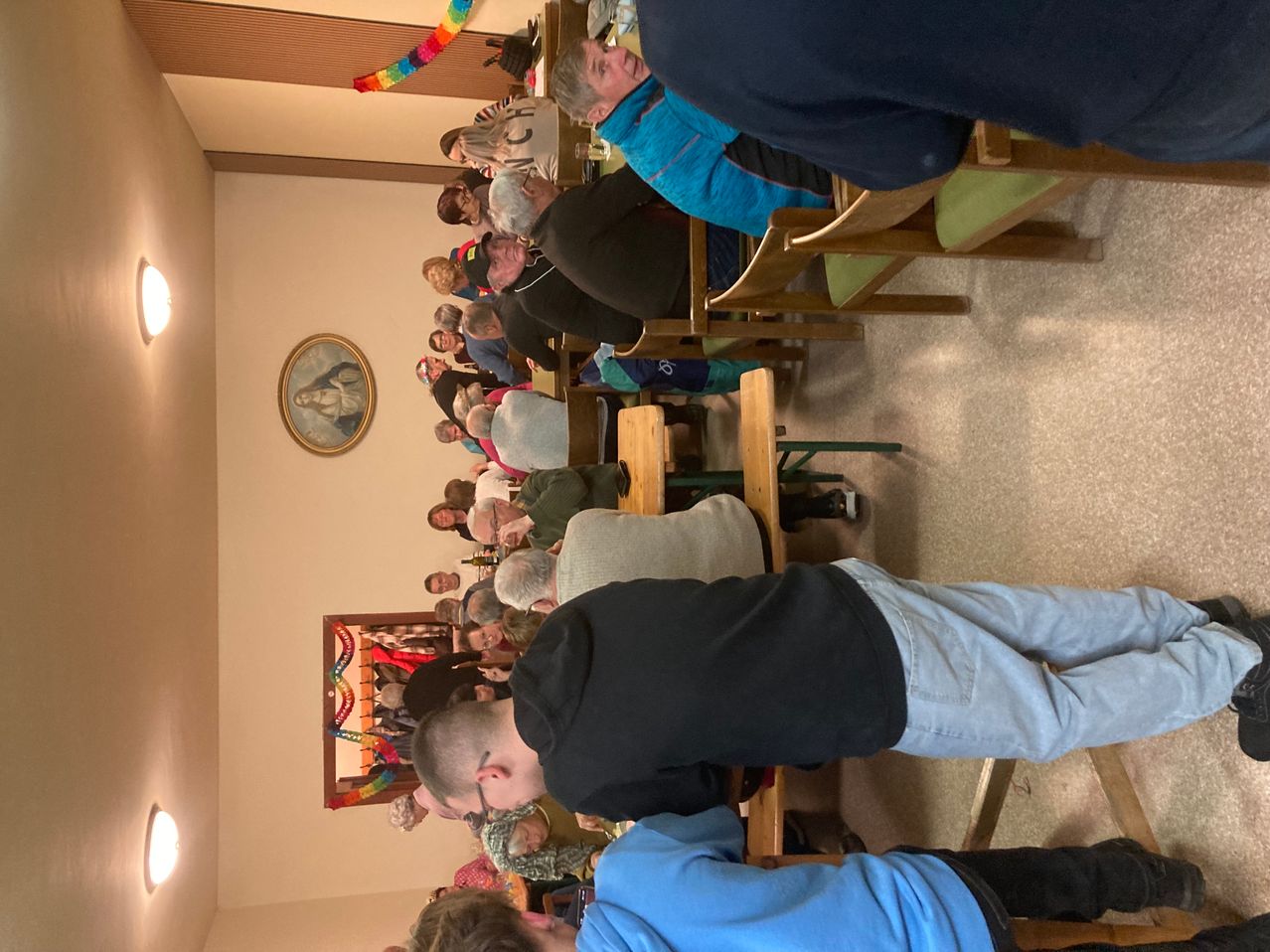 Eine Übersicht von oben auf einen Raum mit vielen Leuten, die um Tische herum sitzen, einige mit dem Rücken gedreht, einem gerahmten Bild an der Wand und bunten Dekorationen.