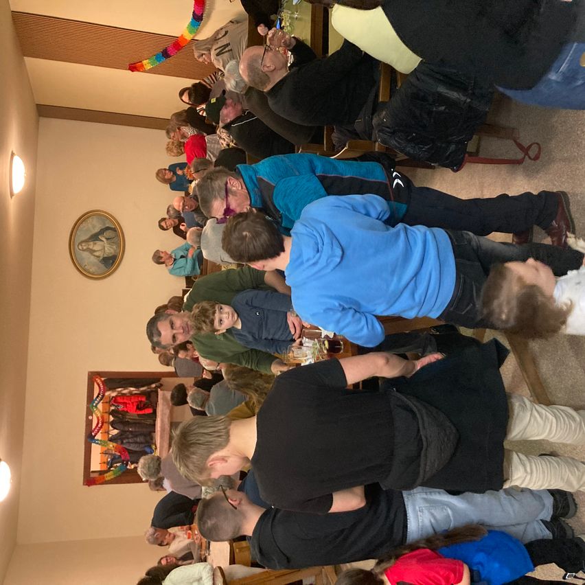 Eine Gruppe von Menschen ist in einem Raum versammelt, wahrscheinlich zu einer Feier. Einige stehen, andere sitzen. Einige tragen Brillen. Ein Mann in einem schwarzen Hemd hält einen Becher. Der Raum hat eine weiße Wand und eine Decke mit Lichtern. Es gibt zwei Gemälde an der Wand.