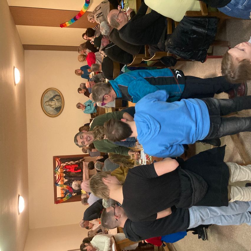 Eine Gruppe von Menschen ist drinnen versammelt, wahrscheinlich in einer Kirche, einige lächeln. Einige tragen Jacken und andere Brillen. Ein Bilderrahmen ist an der Wand montiert.