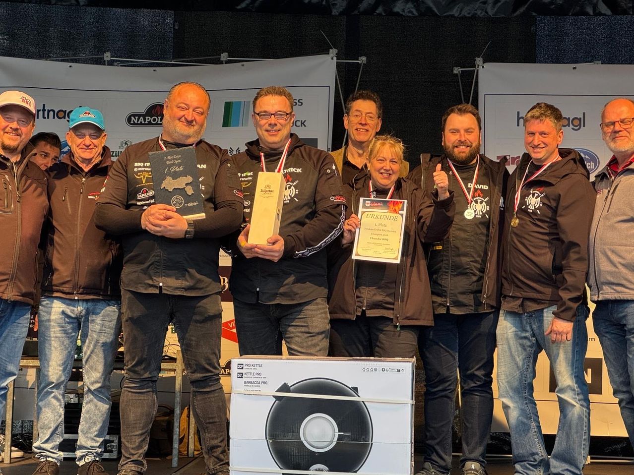 Sechs Männer und eine Frau stehen auf einer Bühne und halten Medaillen und Auszeichnungen. Ein Banner und eine Box sind hinter ihnen. Sie alle lächeln und posieren für ein Foto.