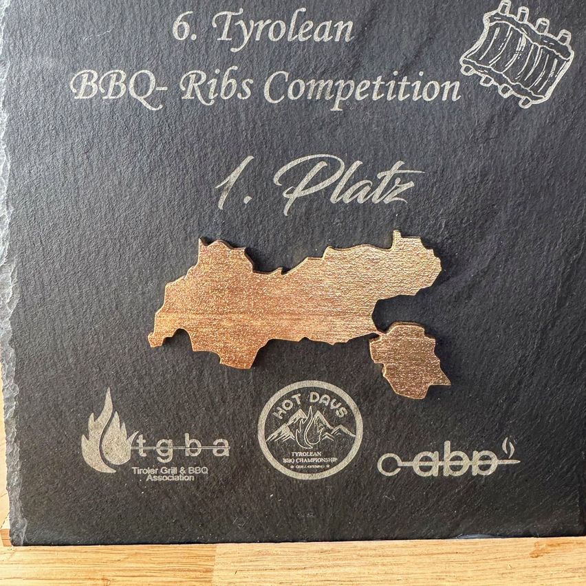 Eine schwarze Plakette mit dem Text 6. Tyrolean BBQ-Ribs Competition 1. Platz, mit einer holzfarbenen Karte von Tirol und Logos für HOT DAYS, tg ba und c-abd.