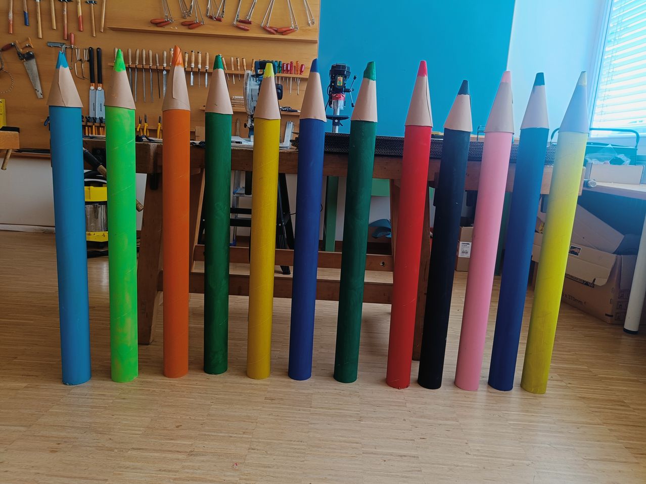 Eine Reihe bunter Stifte, jeder in einer anderen Farbe gestrichen, steht aufrecht auf einem Holzboden. Dahinter befindet sich ein Holztisch mit verschiedenen Werkzeugen und Ausrüstung.