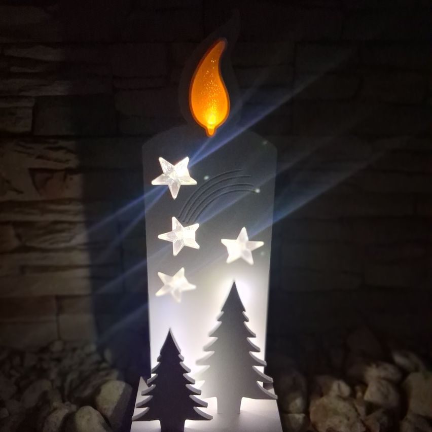 Eine brennende Kerze mit flackernder Flamme, umgeben von Sternen und Bäumen, auf einer felsigen Oberfläche platziert. Der Hintergrund ist dunkel.