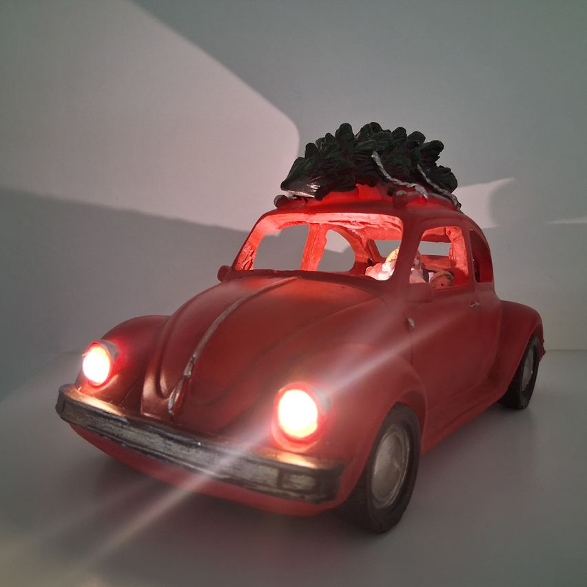 Ein rotes Auto mit einem Weihnachtsbaum oben, eingeschalteten Lichtern und Figuren im Inneren. Das Auto steht auf einer weißen Oberfläche mit Schatten.