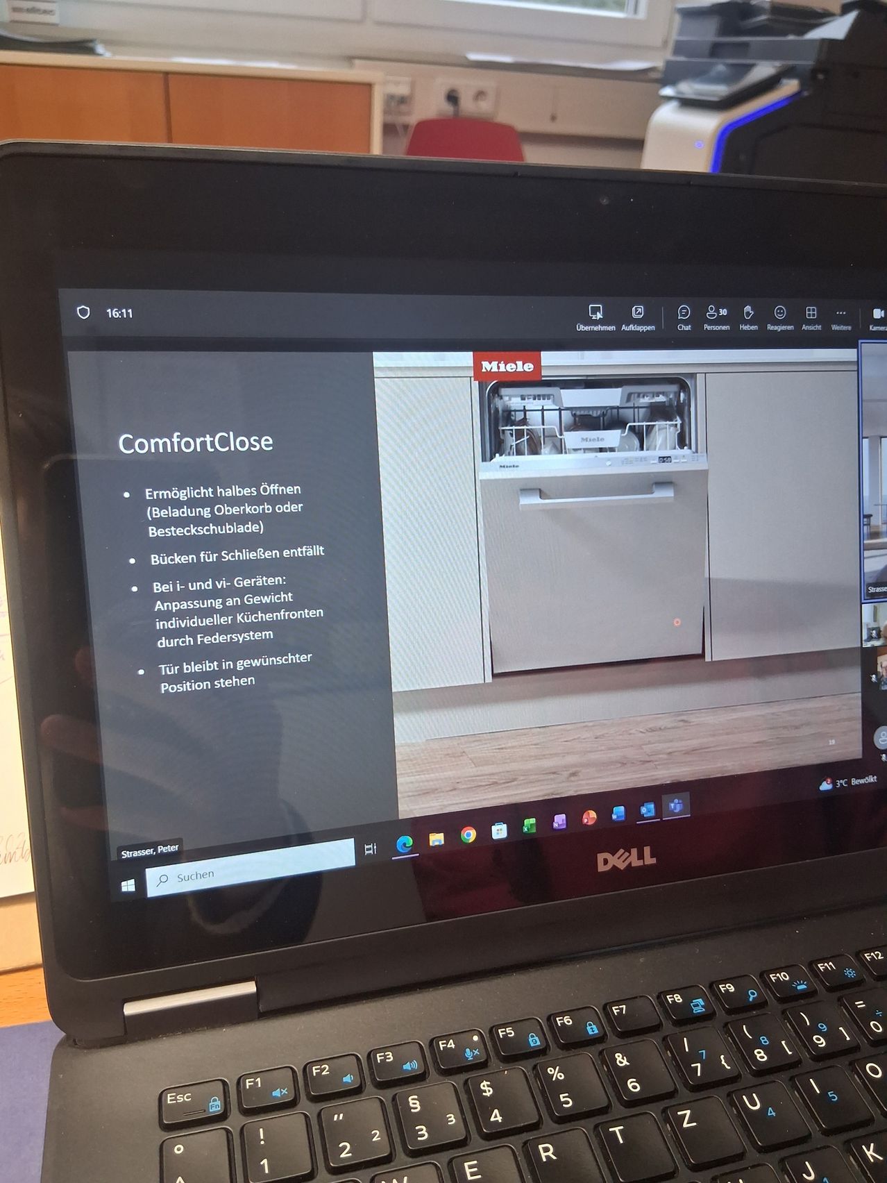 Ein Laptop zeigt einen Bildschirm mit einer Präsentationsfolie über einen Geschirrspüler. Die Folie enthält Text über ComfortClose, das ein teilweises Öffnen, Be- oder Entladen ermöglicht. Es werden auch die Funktionen des Geschirrspülers wie leises Schieben, Gewichtsanpassung und die Tür in der gewünschten Position bleiben erwähnt.