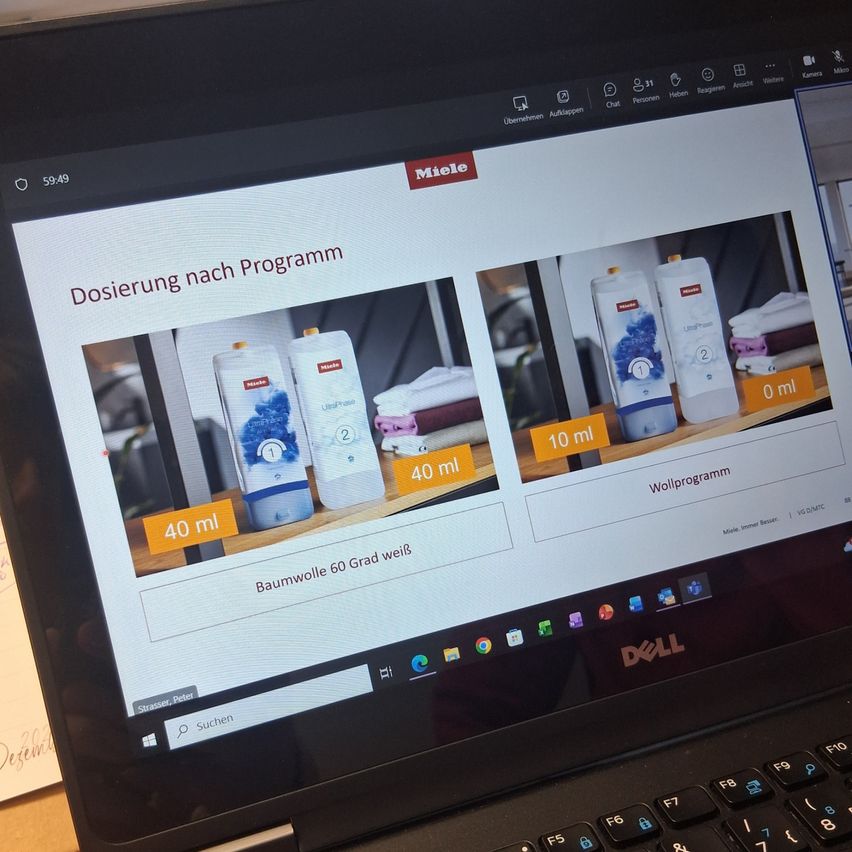Ein Laptop-Bildschirm zeigt eine Mielo-Website-Seite. Es zeigt einen Dosierprogramm-Leitfaden mit Bildern von Waschmittel-Pods. Die Seite zeigt die Dosierung für Baumwolle bei 60 Grad an. Der Bildschirm zeigt auch die Windows-Suchleiste.