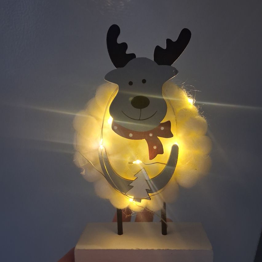 Eine festliche Weihnachtsdekoration eines Rentiers auf einer Wolke mit gelben Lichtern, einem roten Band und einem kleinen Baum. Es steht auf einer weißen Basis vor einem grauen Hintergrund.