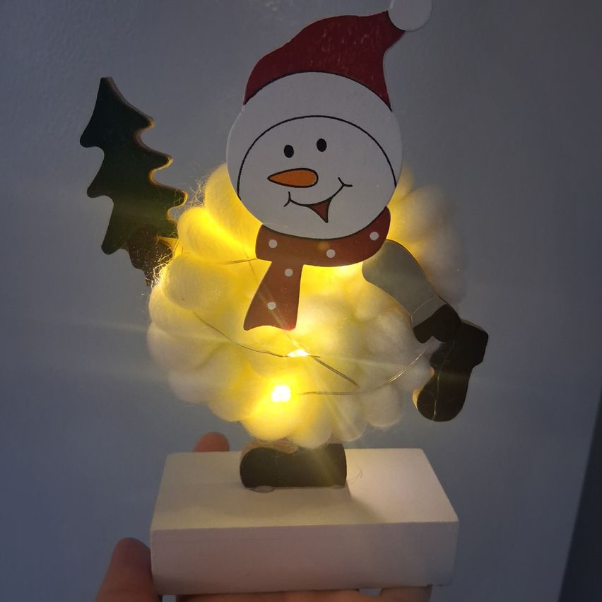 Eine Schneemann-Figur mit einem Weihnachtsmannhut, einer Schürze und gelben Lichtern. Sie steht auf einer weißen Basis und hält einen kleinen grünen Baum.