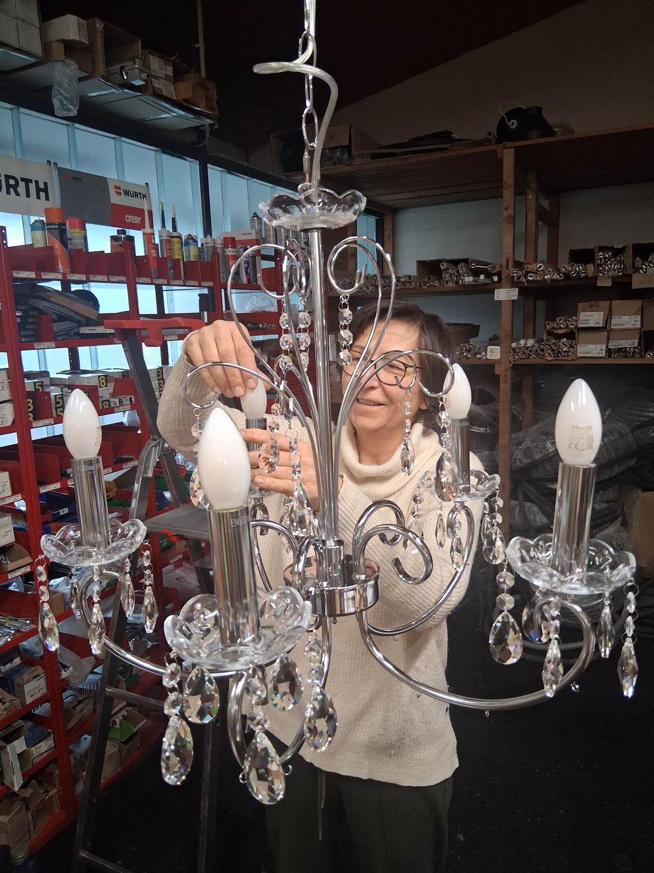 Eine Frau in einem weißen Pullover hält einen Kristallkronleuchter mit sechs Glühbirnen in einer Werkstatt. Hinter ihr befinden sich Regale voller Kisten und Dosen, und ein Schild mit der Aufschrift Wurth.