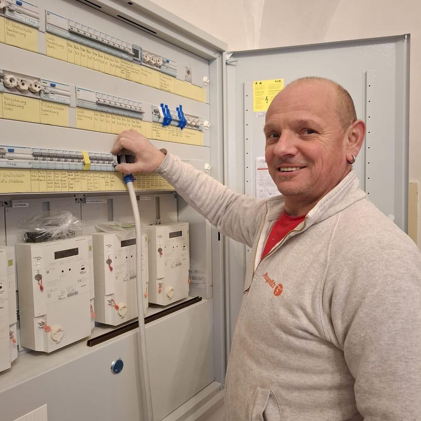 Ein Mann in einer grauen Kapuzenjacke lächelt, während er ein Werkzeug vor einem Schaltschrank mit mehreren Zählern und Schaltern hält.