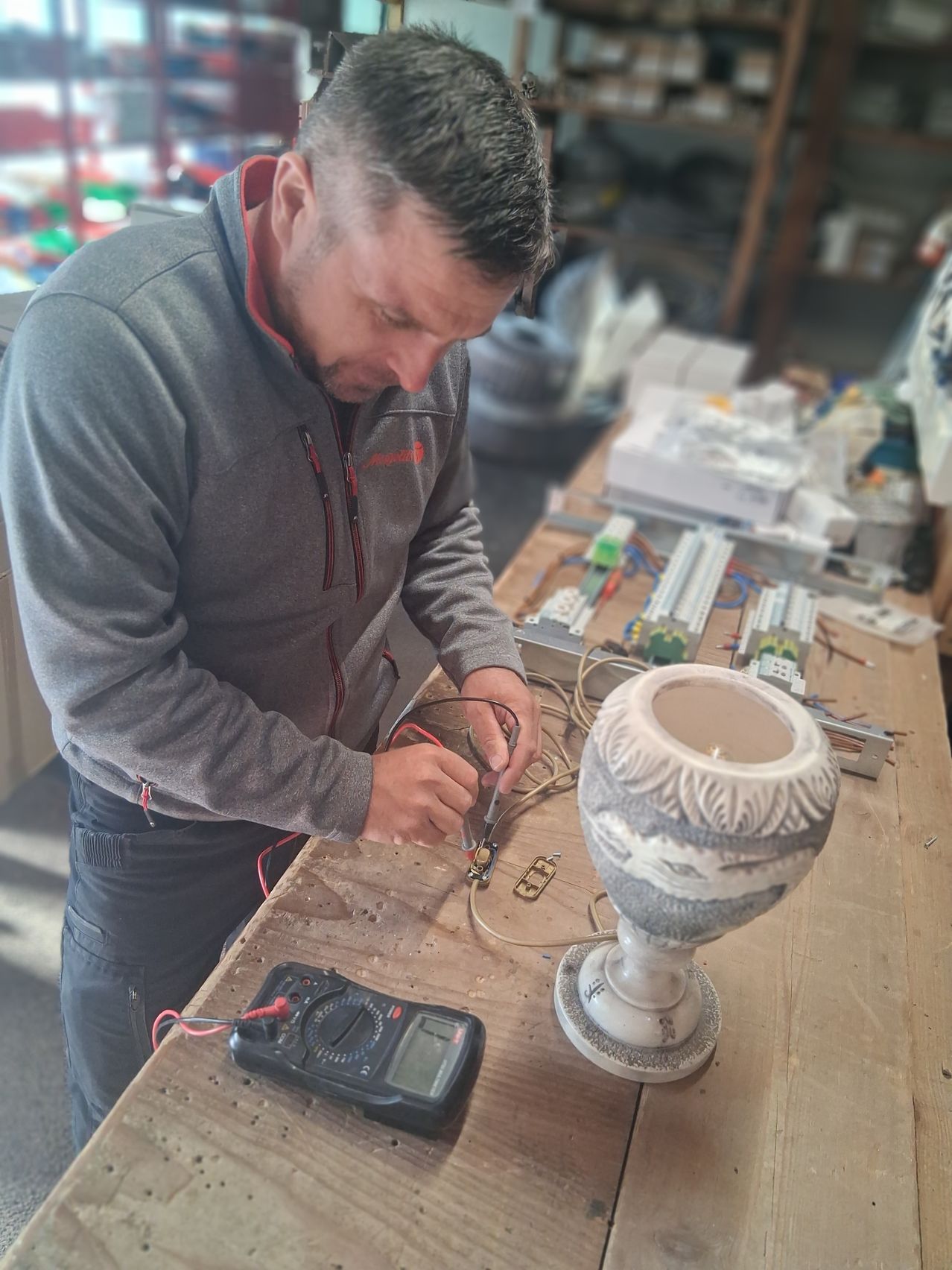 A man is working on a lamp in a workshop, using a multimeter and a soldering iron. The lamp is placed on a wooden table with other electronic components.