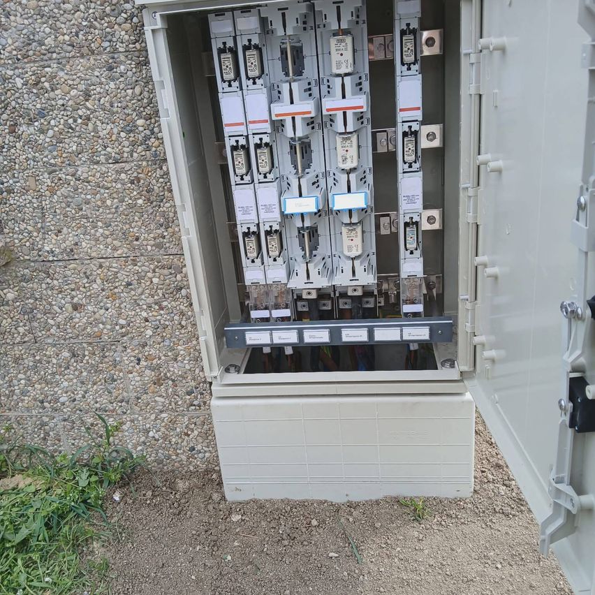 An open electrical distribution panel shows several circuit breakers and switches, positioned against a stone wall.