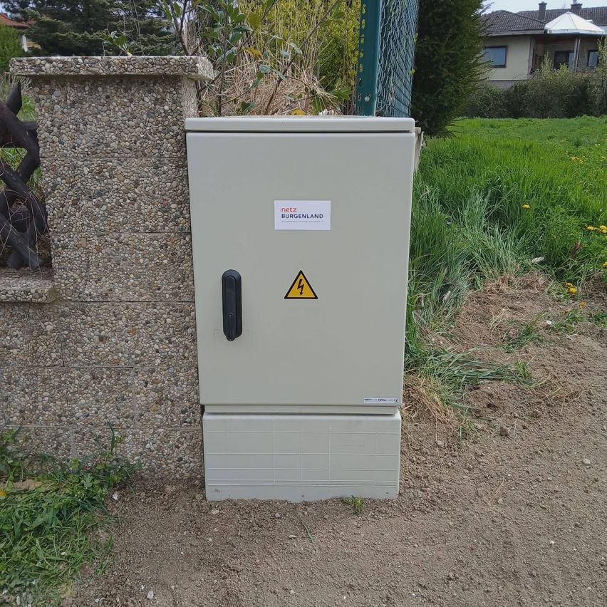 A white electrical box with a yellow warning triangle is fixed to the side of a stone wall near a fence. Behind it, there is a grassy area and a house.