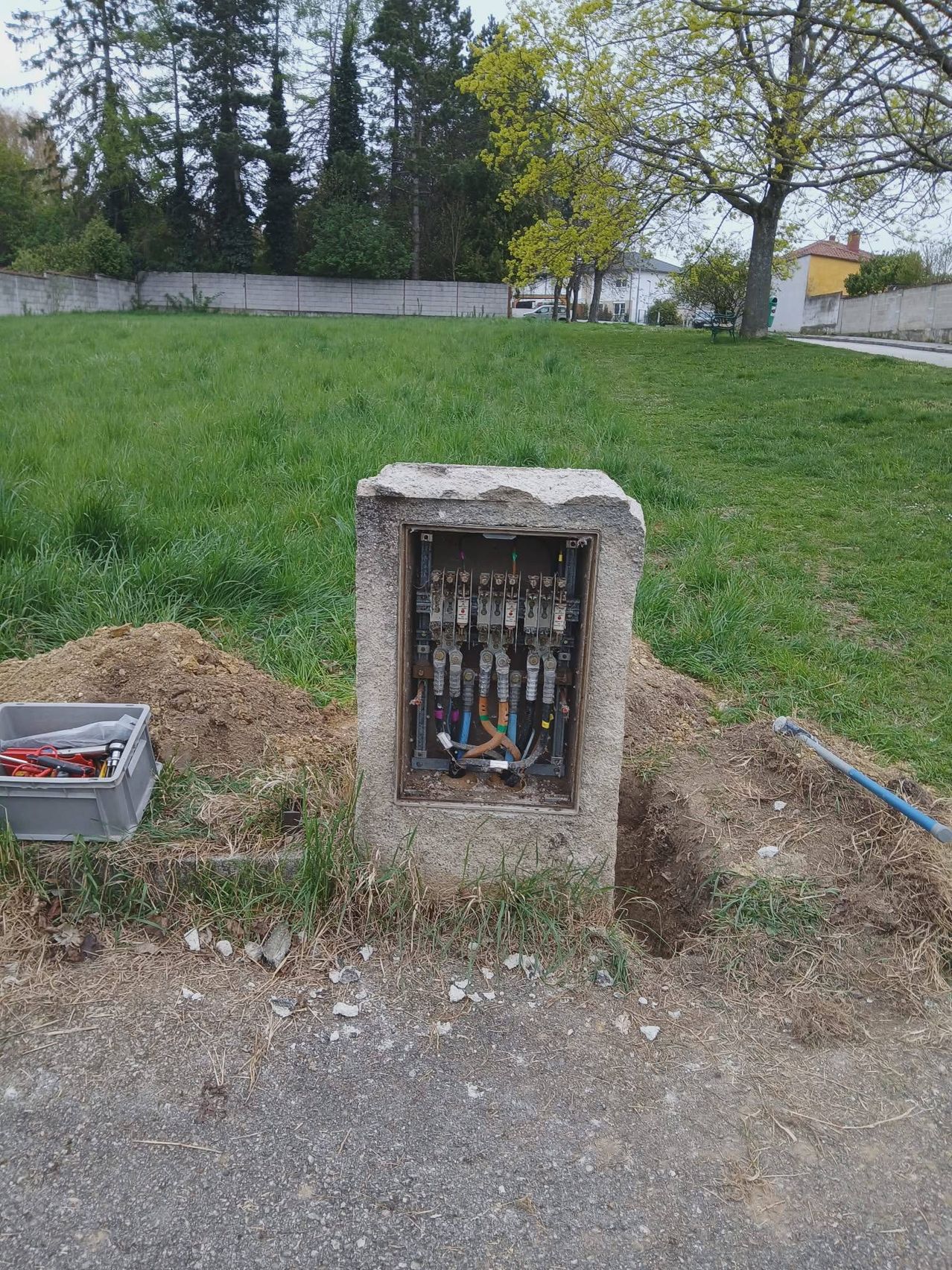 An electrical junction box is buried in a grassy field, with tools and a shovel nearby. A concrete wall and houses are in the distance.
