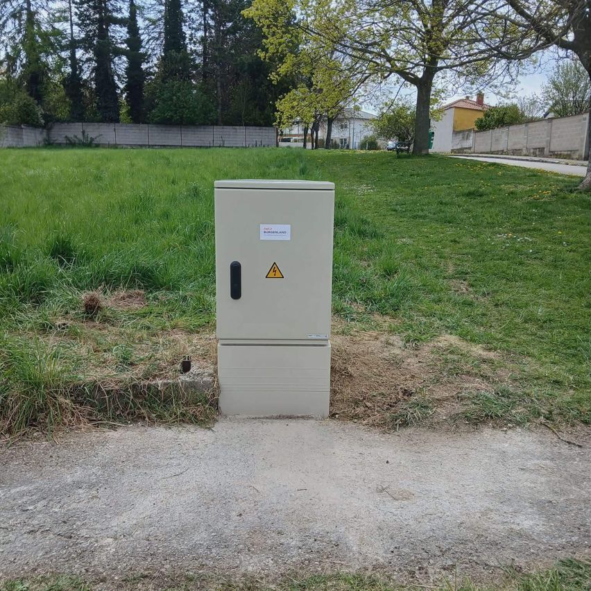 A white electrical box with a yellow caution sign is placed in a grassy field with trees and buildings in the background.