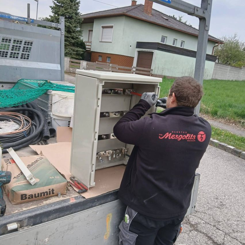 A man works on an electrical box in a truck with a house and yard in the background.