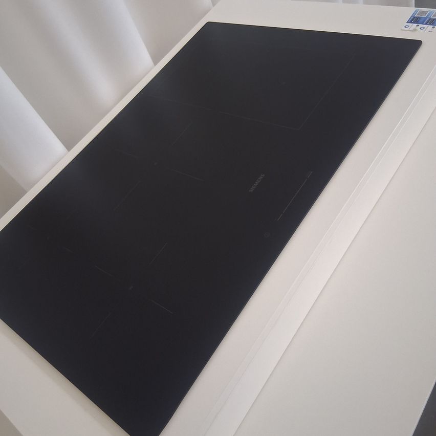 Ein schwarzer Induktionsherd wird auf einer weißen Oberfläche ausgestellt. Er hat vier quadratische Heizzonen und befindet sich noch in der Verpackung.