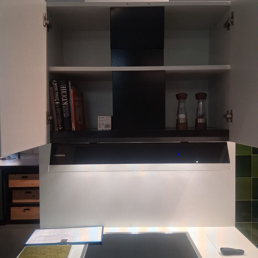 Eine Küche mit weißen Schränken, einer offenen Tür, einem Regal mit Büchern, einer Schachtel und zwei Salz- und Pfefferstreuern, einem Siemens-Gerät und einer Fernbedienung auf einer Arbeitsplatte.