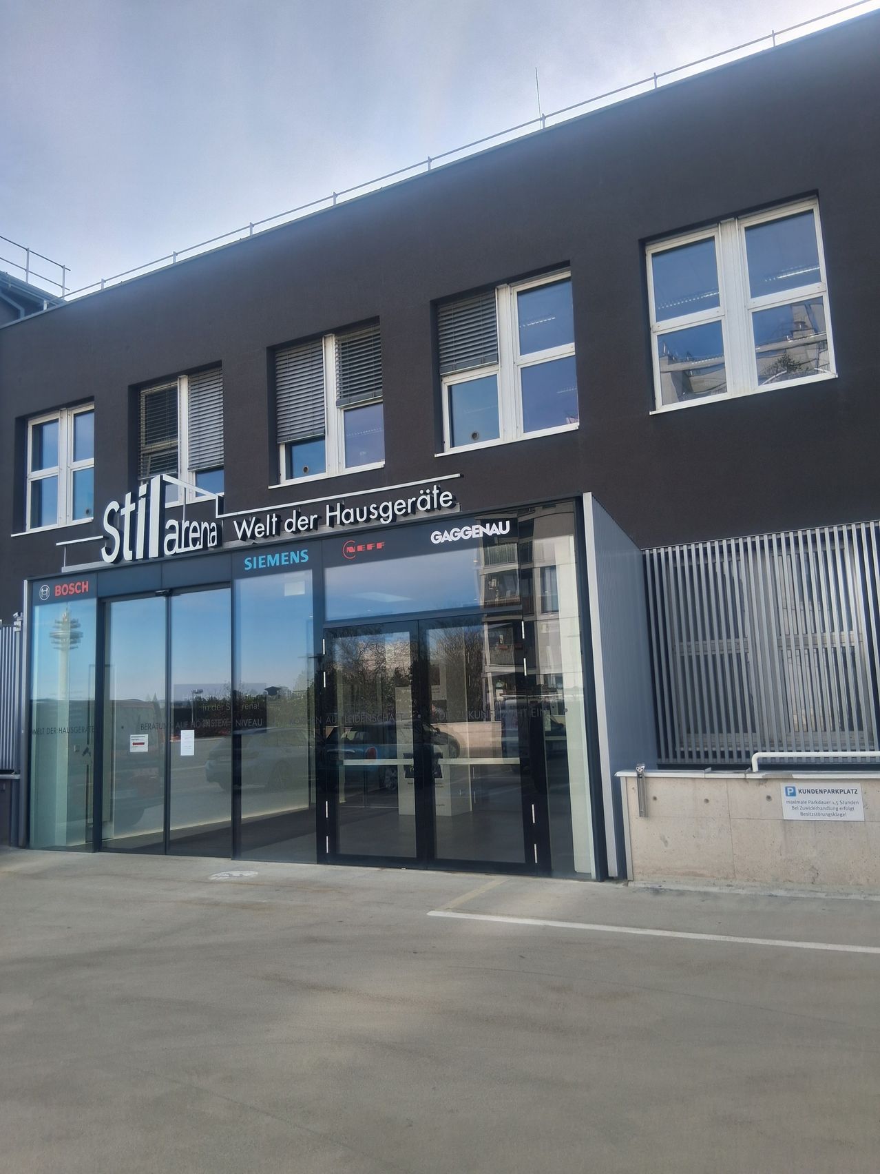 Ein Gebäude mit 'Still arena' Schild, mit Glastüren, Fenstern und Firmenlogos, darunter Siemens, Bosch und Siemens. Der Eingang ist modern mit grauer Außenseite.