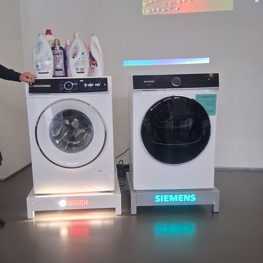Zwei Waschmaschinen, eine von Bosch und eine von Siemens, sind mit Waschmittelflaschen darauf ausgestellt. Sie haben beleuchtete Logos und stehen auf einem grauen Boden.