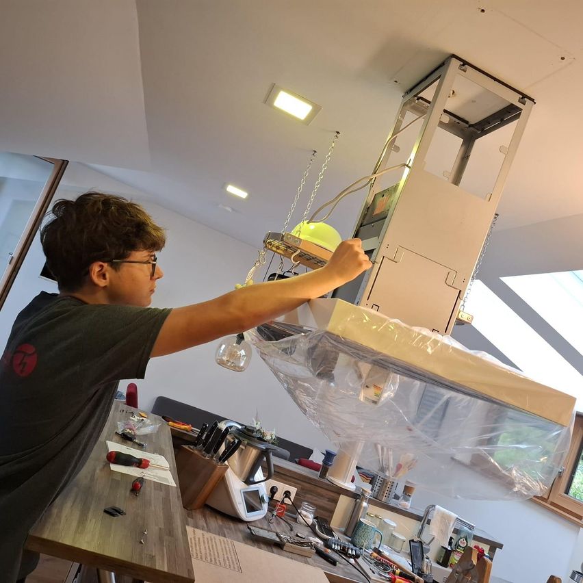 A young man is working on an appliance, possibly a kitchen extractor fan, with a plastic cover on it. He is holding a tool and seems to be fixing it. There are other tools and appliances on the table in front of him.