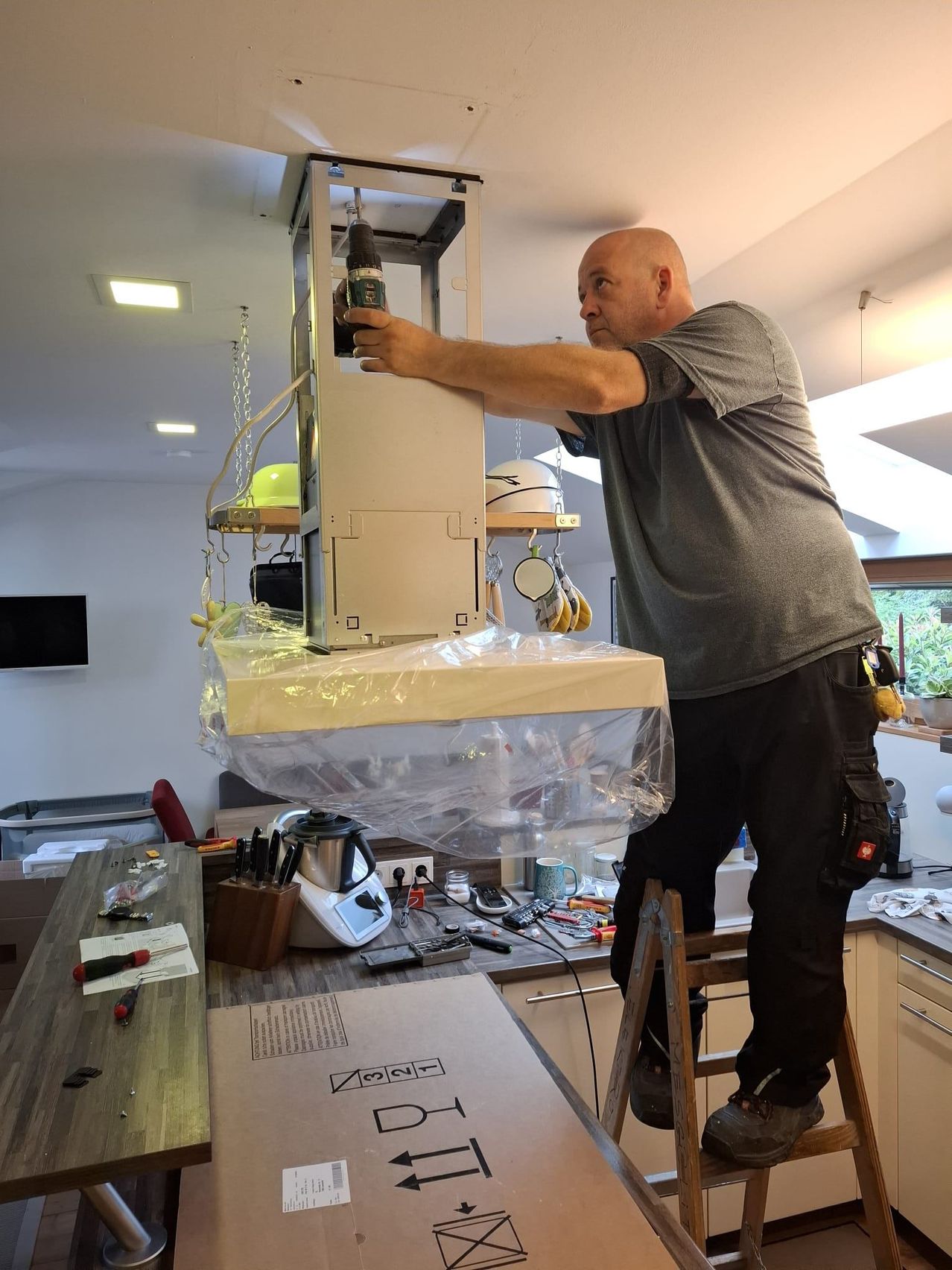 A man is working on a kitchen appliance, possibly a blender, while standing on a stool. He is holding a tool and appears to be fixing or assembling it. The kitchen has various items, including a trash bin, a TV, and a countertop with a coffee machine.
