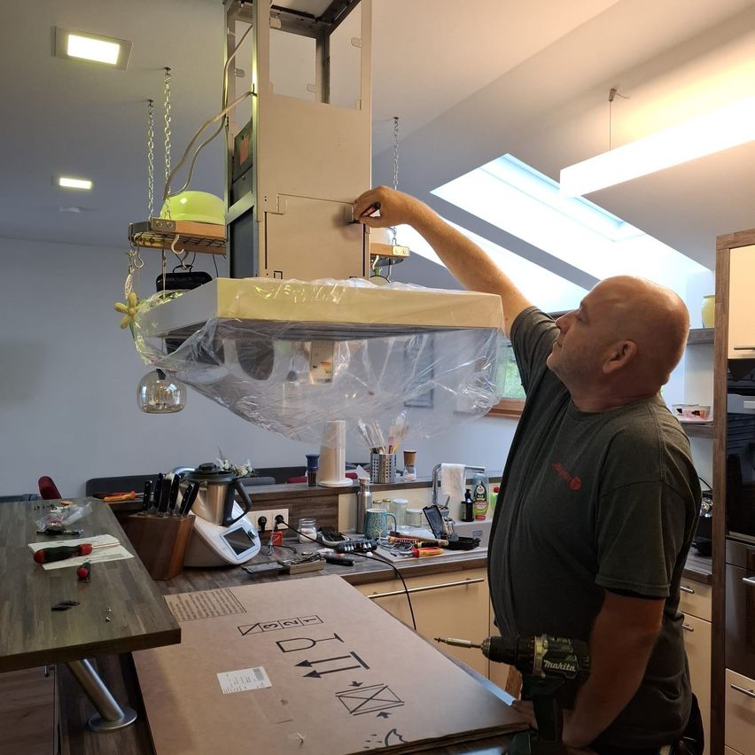 A man is installing a kitchen hood with a plastic cover over it. He is holding a drill and has a toolbox on the table.