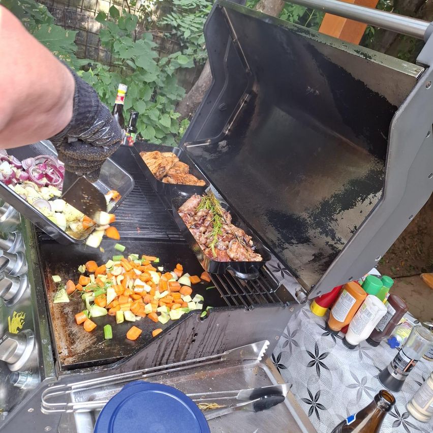 A person wearing gloves is grilling meat and vegetables on an outdoor grill. Various vegetables and meat are on the grill, and bottles of sauces are on the table.