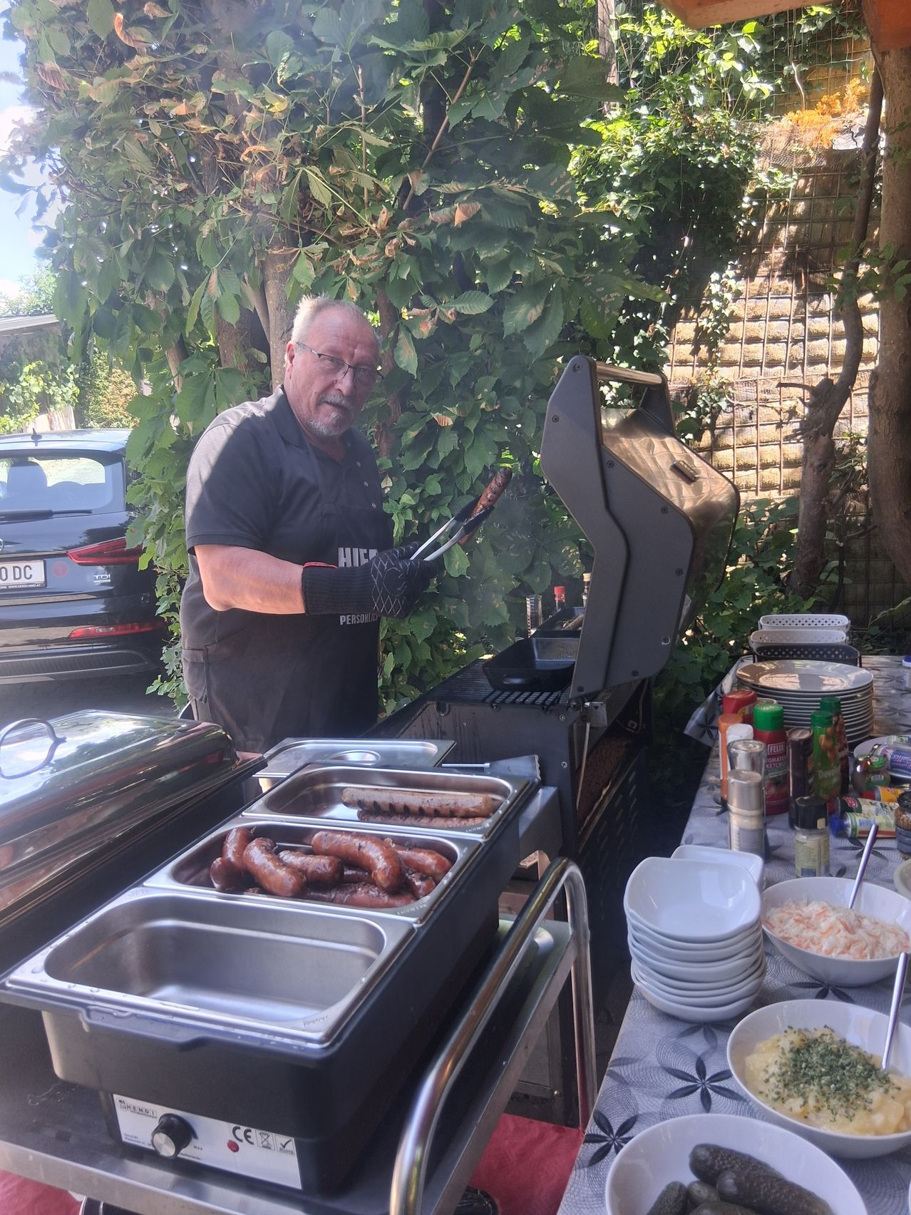 A man is grilling sausages outdoors. He is wearing gloves and standing next to a table with plates, bowls, and condiments.