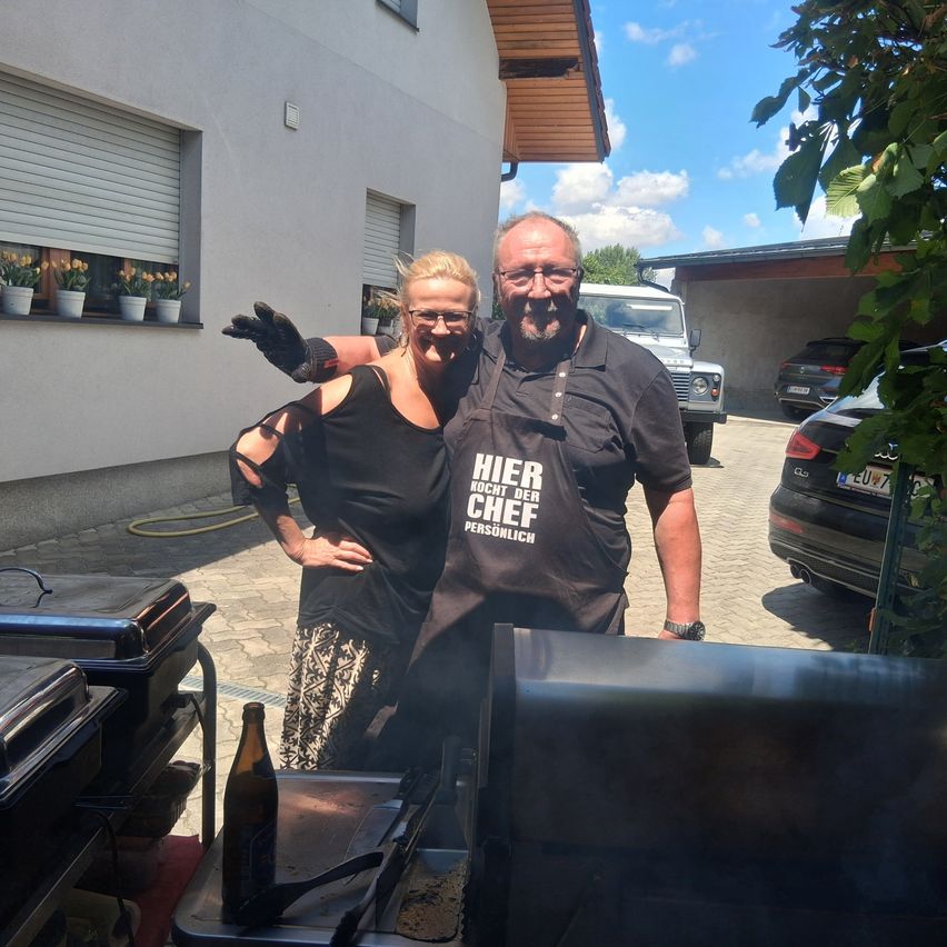 A man and a woman stand in front of a grill, wearing black aprons with the words 'Hier kocht der Chef persönlich.' They smile, with the woman holding the man's arm.