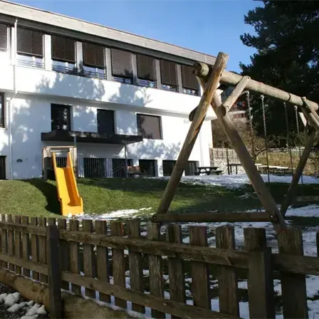 Ein weißes Gebäude mit mehreren Fenstern und einem gelben Rutschen davor, umgeben von einem Holzzaun und Schnee auf dem Boden.