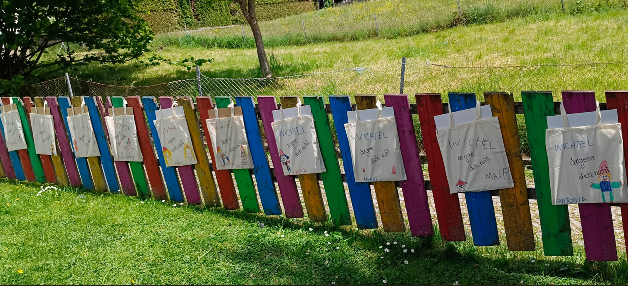 A colorful fence with bags labeled 'WICHTEL' and 'HENRY' hangs from it, in a grassy field with a tree and wire fence in the background.