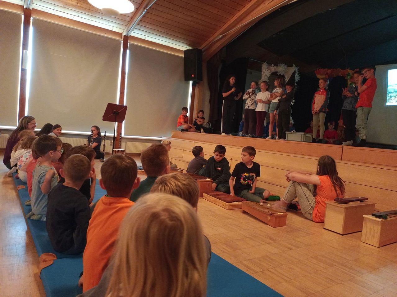 Kinder sitzen auf dem Boden in einem Klassenzimmer und schauen sich eine Aufführung auf der Bühne an. Einige Kinder spielen Xylophon.