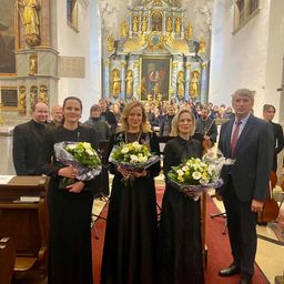 Vier Personen stehen vor einem Kirchenaltar, zwei halten Blumensträuße. Dahinter spielt eine Gruppe von Musikern im Hintergrund Instrumente.