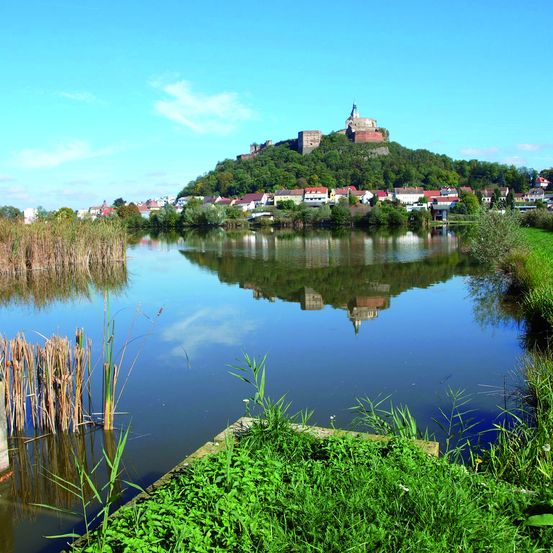 Eine Burg steht auf einem Hügel über einem Fluss und einer Stadt. Die Burg spiegelt sich im Wasser, und der Himmel ist klar und blau.
